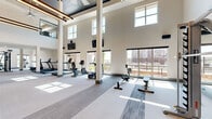 Located on N Oklahoma Avenue in Oklahoma City, this generously sized fitness center offers an impressive and well-appointed exercise facility across a single open floor. The space immediately commands attention with its soaring double-height ceilings, accentuated by large clerestory windows that flood the interior with abundant natural light throughout the day. Structural columns are elegantly integrated into the layout, complementing the modern, neutral color palette of white walls, soft gray carpeting with contrasting linear carpet tile accents, and teal-toned column bases.

The main exercise area is thoughtfully organized to accommodate a wide range of fitness activities. A dedicated cardio zone lines the windowed perimeter, housing treadmills, ellipticals, stationary bikes, and rowing machines, all oriented toward expansive views of the surrounding community. Strength training equipment, including a multi-station cable machine and free weight benches, is positioned throughout the open floor plan, providing ample room for movement.

A striking industrial-style staircase with a metal railing ascends one end of the space, hinting at additional access points above. Mounted flat-screen televisions and ceiling fans with pendant lighting add both functionality and visual appeal. A lower-ceilinged secondary zone extends the footprint further, offering additional workout space and utility areas. The overall ambiance is modern, airy, and motivating.