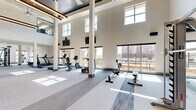 Located on N Oklahoma Avenue in Oklahoma City, this generously sized fitness center offers an impressive and well-appointed exercise facility across a single open floor. The space immediately commands attention with its soaring double-height ceilings, accentuated by large clerestory windows that flood the interior with abundant natural light throughout the day. Structural columns are elegantly integrated into the layout, complementing the modern, neutral color palette of white walls, soft gray carpeting with contrasting linear carpet tile accents, and teal-toned column bases.

The main exercise area is thoughtfully organized to accommodate a wide range of fitness activities. A dedicated cardio zone lines the windowed perimeter, housing treadmills, ellipticals, stationary bikes, and rowing machines, all oriented toward expansive views of the surrounding community. Strength training equipment, including a multi-station cable machine and free weight benches, is positioned throughout the open floor plan, providing ample room for movement.

A striking industrial-style staircase with a metal railing ascends one end of the space, hinting at additional access points above. Mounted flat-screen televisions and ceiling fans with pendant lighting add both functionality and visual appeal. A lower-ceilinged secondary zone extends the footprint further, offering additional workout space and utility areas. The overall ambiance is modern, airy, and motivating.