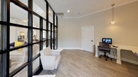 This contemporary single-floor office space makes an immediate impression with its signature floor-to-ceiling black steel-framed glass partition walls, which elegantly divide the interior while maintaining an open, connected feel throughout. The design strikes a refined balance between modern aesthetics and functional practicality.

The reception and waiting area is bathed in abundant natural light streaming through large windows and the glass partitions, creating a bright and welcoming atmosphere. Neutral-toned upholstered lounge chairs and accent seating are arranged to provide a comfortable space for visitors, complemented by light hardwood flooring that runs consistently throughout.

Adjacent to the waiting area, a dedicated workstation zone features built-in white desks with multiple computer workstations, a printer, and ample workspace for staff. Pendant lighting hangs above the desk area, adding a warm, focused glow alongside recessed ceiling lights. Crown molding and clean white trim details lend a polished, professional finish to the space.

The overall ambiance is sophisticated yet approachable — neutral gray and white tones on the walls pair seamlessly with the bold black framing of the glass partitions, creating a visually striking contrast. Outdoor views visible through the windows further enhance the sense of openness, making this a bright and productive professional environment.