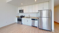 This single-floor space offers a thoughtfully designed layout with a clean, contemporary aesthetic throughout. The living area is a standout feature, boasting soaring ceilings and a trio of tall arched windows with dark frames that flood the room with natural light and offer views of surrounding greenery. Light wood-toned flooring extends throughout the main living areas, contributing to a warm, cohesive feel.

The kitchen is efficiently organized along a single wall and is outfitted with a full suite of stainless steel appliances, including a range, over-the-range microwave, dishwasher, and refrigerator. White shaker-style cabinetry extends to the ceiling, maximizing storage, while a neutral countertop and recessed lighting complete the functional workspace.

The bedroom is a well-proportioned, quiet retreat featuring plush wall-to-wall carpeting and crisp white walls, offering a comfortable contrast to the hard flooring elsewhere. A separate room with wood-look flooring and a large arched window provides additional flexible living space with ample natural light.

The bathroom is clean and functional, featuring a white vanity with warm-toned countertops, a mirrored medicine cabinet, and a glass-enclosed walk-in shower. Light wood-toned flooring ties the bathroom's design to the rest of the unit. Overall, the space presents a bright, modern, and move-in ready environment.
