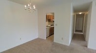 This single-story space offers a clean, functional layout with clearly defined living areas. The dining area is finished with neutral wall-to-wall carpeting and features an elegant chandelier with candle-style lights, adding a classic touch to the otherwise simple, understated design. The walls are painted in a crisp, neutral white, creating a bright and open atmosphere throughout.

An open doorway connects the dining area directly to the kitchen, which features warm-toned wooden cabinetry arranged along the walls, providing ample storage. The kitchen is equipped with a white dishwasher, a sink, and overhead cabinets that extend to a range hood, offering a practical and well-organized cooking space. The kitchen floor transitions from carpet to a hard surface, helping to delineate the two areas. The layout promotes easy flow between spaces, making the overall footprint feel efficient and accessible. The neutral palette and simple finishes throughout give the space a versatile foundation, well-suited for a variety of furnishing styles and personal touches.