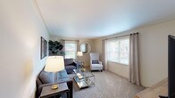This single-floor residence offers a thoughtfully designed layout that balances comfort and functionality across its key living spaces. The living room is generously sized, featuring plush wall-to-wall carpeting, neutral-toned walls, and multiple windows dressed with grommet-panel curtains that invite abundant natural light. A round decorative mirror and framed botanical artwork add character to the space, which flows seamlessly between a sectional seating arrangement and a dedicated media area with an open shelving entertainment unit. A compact dining nook with dark hardwood flooring provides an intimate setting for meals, featuring a round pedestal table flanked by cross-back chairs beneath a sculptural metal wall accent. The kitchen is well-equipped with white cabinetry, dark laminate countertops, stainless steel appliances including a gas range with overhead microwave, and a single-basin sink — all finished with a practical hanging wine glass rack. Both bedrooms are carpeted and serene, each benefiting from natural light through curtained windows. The primary bedroom is notably spacious, offering a walk-in closet and ample room for substantial furniture. The second bedroom mirrors the calm aesthetic with dark wood furnishings and soft, neutral tones. Overall, the residence presents a clean, cohesive design with a warm and livable ambiance throughout.