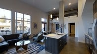 This single-floor loft-style space showcases an impressive open-concept layout defined by its dramatic ceiling height, which creates an expansive, airy atmosphere throughout. A prominent exposed concrete column anchors the central living area, lending an industrial character that complements the overall design aesthetic.

The kitchen is a standout feature, centered around a large dark-cabinetry island with a crisp white countertop and an integrated sink. Pendant lighting hangs above the island, adding both functional illumination and a refined visual element. The polished concrete floors extend seamlessly throughout the space, reinforcing the industrial-modern style.

The living area is bathed in natural light streaming through large windows along the exterior wall, offering views of the surrounding neighborhood. A leaning ladder leads to an upper loft or mezzanine level, adding vertical interest and additional functional space. A barn-style sliding door provides a rustic contrast to the otherwise contemporary design.

The open dining and living zones flow naturally from the kitchen, creating a cohesive, connected layout ideal for both everyday living and entertaining. The overall ambiance balances industrial elements with modern finishes for a sophisticated, urban feel.