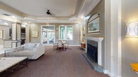 Located at 1060 Coral Ridge Drive in Coral Springs, Florida, this single-story clubhouse offers a sophisticated and welcoming environment designed for community gathering and leisure. The main lounge serves as the heart of the facility, featuring a stately marble fireplace flanked by decorative arched niches, tray ceilings with crown molding, and plush carpeting with an ornate pattern. Double glass-paned entry doors flood the space with natural light, complementing the elegant seating arrangements and framed artwork throughout.

Adjacent to the main lounge is a cozy media room outfitted with a built-in hardwood entertainment center, arched display shelving, and large windows dressed with warm valances and wooden blinds that filter natural light beautifully. A comfortable sofa and glass-top coffee table complete this inviting retreat.

The billiards room provides a spirited recreational space, centered around a full-size pool table beneath a stylish pendant light fixture. Tray ceilings, two-tone wall paneling in soft teal, and a French door opening to greenery outside lend the room a bright, airy character.

A dedicated card room offers intimate seating around glass-topped tables, adorned with botanical artwork and warm earth-tone walls. The clubhouse also includes a well-appointed kitchenette featuring dark cabinetry, granite countertops, stainless steel appliances, and a pass-through window overlooking the corridor — ideal for catering and events.