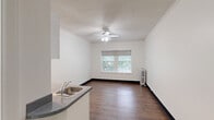 Located at 1410 SW Broadway in Portland, Oregon, this efficient studio unit offers a well-organized single-floor layout that makes practical use of every square foot. The main living area features warm hardwood-style flooring, crisp white walls, and a textured ceiling with a central ceiling fan that promotes air circulation and adds a functional focal point to the room. A large double-paned window fitted with a roller shade allows natural light to fill the space while offering pleasant views of surrounding greenery. A classic cast-iron radiator provides supplemental heating, adding a touch of vintage character. Crown molding runs along the upper perimeter of the main room, lending a subtle architectural detail that elevates the overall finish. The kitchen area is neatly appointed with white cabinetry, granite-look countertops, a stainless steel sink, a compact refrigerator, and a white electric range — providing all the essential cooking amenities in a streamlined footprint. The bathroom includes a full tub and shower combination enclosed by clean white subway-style tile, maintaining a simple and functional aesthetic. Throughout the unit, the consistent use of neutral tones and wood-look flooring creates a cohesive and tidy living environment suited for efficient urban living in the heart of Portland.