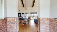 Located at 6205 Longstem Way in Loveland, Colorado, this single-story community space at Vella Terra offers a thoughtfully designed interior that blends rustic character with modern style. The open floor plan is anchored by rich cork flooring throughout, complemented by exposed brick wainscoting and soaring vaulted ceilings that lend a sense of grand, airy openness.

The kitchen is a standout feature, showcasing slate-gray shaker cabinetry, a mint-green central island with a stone countertop, a dramatic brick archway housing a statement range hood, and open wood shelving. A sliding barn door adds a rustic-industrial accent. The space flows seamlessly into the dining area, which features wood-topped tables, upholstered seating, built-in bench banquettes, and a curated built-in cabinet wall with a chalkboard focal point and flanking bookshelves — ideal for gatherings or community events.

The living lounge is bathed in natural light from large windows overlooking the surrounding neighborhood and open skies. Sage-green walls, floating wood shelves, and eclectic furnishings including a live-edge coffee table and plush blue sofa create a warm, inviting atmosphere. The entrance area connects to outdoor spaces with views of a pool area, completing this vibrant community hub.