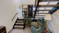This space makes a strong architectural statement with its open-tread staircase as a central design feature. The staircase, constructed with rustic wooden treads and a sleek black metal frame and railing, draws the eye upward and serves as both a functional and visually striking element. The entry area is anchored by a bold black door framed by windows on either side, allowing natural light to filter into the space and create a bright, welcoming atmosphere.

The lower living area is carpeted and designed for comfort, featuring a plush teal upholstered recliner accented with a decorative pillow, positioned near a potted indoor plant that adds a touch of greenery. A baseboard heater runs along the wall near the entryway, ensuring warmth throughout the space. The walls are finished in a clean, neutral tone, complementing the mix of rustic and industrial design elements.

A wall-mounted notice board near the entry adds a practical touch. The overall ambiance balances a cozy, lived-in character with distinctive architectural detailing, making this space both functional and visually interesting.