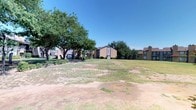 This residential complex presents a well-established community characterized by its classic brick construction and sprawling outdoor common areas. The grounds are defined by generous open lawns that stretch between the buildings, offering residents ample shared outdoor space. Mature trees are distributed throughout the property, providing natural shade and contributing to a settled, established atmosphere.

The multi-unit buildings feature traditional pitched rooflines with chimneys, brick and neutral-toned exteriors, and covered balconies or patios on upper and lower levels — adding architectural dimension while offering private outdoor access for residents. Trimmed hedges and modest landscaping line the building entrances, adding a measure of greenery and structure to the grounds.

The single-story community center or leasing building is a low-profile structure set amid the greenery, featuring a wide roofline and neatly maintained surrounding hedges. The overall layout of the community feels open and spacious, with clear sightlines across the lawns from multiple vantage points. The property's design reflects a practical, community-oriented approach, balancing outdoor recreational space with residential density in a setting that evokes a calm, suburban character beneath clear, expansive skies.