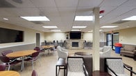 This single-floor communal space is thoughtfully designed to accommodate a variety of social settings, combining comfort and functionality in an open, well-lit layout. The room is anchored by a striking corner stone fireplace, which serves as a natural focal point and lends a warm, inviting character to the space. A large flat-screen television is mounted above the fireplace, making it a natural hub for entertainment and group viewing.

The space is divided into distinct seating zones to support different activities. A lounge area near the fireplace features upholstered armchairs with patterned fabric arranged around small side tables, creating an intimate setting for conversation. On the opposite side, café-style round tables paired with standard dining chairs provide a more structured environment suitable for meals or collaborative work. High-top tables with bar-height seating along the perimeter offer additional flexible options.

Overhead, a drop-panel ceiling with evenly spaced recessed fluorescent lighting ensures the entire room is consistently and brightly illuminated. The neutral wall tones and light tile flooring contribute to a clean, open atmosphere. Structural columns are integrated naturally into the layout without interrupting the flow of the space. Overall, the room balances practicality with a comfortable, community-oriented ambiance.