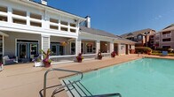 Located at 8531 Summersweet Ln in Raleigh, NC, this community amenity space is designed to deliver a resort-like outdoor living experience. The centerpiece is a generously sized swimming pool with crystal-clear turquoise water, complemented by a surrounding concrete deck outfitted with lounge chairs and seating areas perfect for relaxing under the North Carolina sun. A pergola with additional outdoor seating offers a shaded retreat within the landscaped grounds, which feature mature trees, colorful flowering plants, and well-maintained hedgerows that create a sense of privacy and natural beauty. The single-story clubhouse building is constructed in a classic Southern architectural style, with horizontal lap siding, a gabled roofline, and prominent chimneys. A covered veranda stretches along the building's poolside façade, supported by stately white columns and adorned with hanging flower baskets. The veranda is furnished with plush wicker seating, making it an inviting space for socializing or unwinding while overlooking the pool. Large windows and French doors connect the interior to the outdoor environment, enhancing the flow between inside and outside spaces. The overall ambiance is welcoming, vibrant, and thoughtfully landscaped, offering residents a well-appointed outdoor destination within the community.