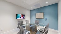 Located at 5961 SW 68th St in South Miami, FL, this compact conference room is thoughtfully designed to maximize functionality within an intimate footprint. The room features a modern glass-top conference table with a wood-toned base, surrounded by six ergonomic mesh chairs that provide both comfort and a professional aesthetic. The interior design is defined by a crisp, neutral palette of white and light gray, punctuated by a bold teal accent wall that adds depth and visual interest to the space. Recessed lighting throughout ensures consistent, well-distributed illumination across the entire room. Two wall-mounted flat-screen displays offer versatile presentation capabilities, while a whiteboard on the teal accent wall provides a space for collaborative note-taking and brainstorming. A wall-mounted control panel allows for convenient room management. Light-toned tile flooring runs throughout, contributing to the clean and polished atmosphere. Large glass-paneled doors open to an adjacent, brightly lit area, allowing for a sense of connectivity to the broader office environment. Framed artwork accents the walls, adding a refined touch to the professional setting. This conference room is well-suited for small team meetings, presentations, and focused collaborative sessions.