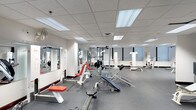 This fitness center is designed to support a full range of exercise routines in a clean, functional environment. The space features a drop ceiling with recessed fluorescent panel lighting that evenly illuminates the entire floor, creating a bright and energizing atmosphere conducive to focused workouts. The flooring consists of durable rubber gym matting in a neutral gray tone, providing traction and impact absorption throughout the area.

The room is outfitted with a diverse selection of strength training equipment, including adjustable weight benches, barbell racks, cable machines, and various resistance stations. Equipment is arranged with ample spacing to allow users to move freely between stations. Along one wall, large mirrors are mounted from mid-height, extending across a significant portion of the room, allowing users to monitor their form during exercises.

The layout is open and spacious, supporting simultaneous use by multiple individuals without a sense of crowding. The white walls and light-toned ceiling contribute to the room's clean, professional aesthetic, making this fitness center a well-appointed facility suited for both casual and dedicated training.