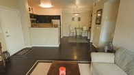 This single-floor residence offers an open-concept design that seamlessly connects the kitchen, dining, and living areas into one cohesive, flowing space. Dark hardwood floors run throughout, providing a rich contrast against the warm, neutral-toned walls and creating a sense of grounded elegance.

The kitchen is positioned at the rear of the open layout and features dark cabinetry that extends to the upper walls, light-toned countertops, and essential appliances including a microwave and refrigerator. A raised breakfast bar separates the kitchen from the rest of the living space, offering both a functional workspace and a casual dining option.

Adjacent to the kitchen, a compact dining area accommodates a glass-top bistro table with sleek metal chairs, illuminated by a pendant light fixture that adds a modern, refined touch. Built-in shelving nearby provides display and storage space.

The living area is defined by a neutral area rug and is furnished with a comfortable upholstered chair, anchored by a dark coffee table. Warm overhead lighting enhances the cozy ambiance throughout. A hallway at the rear leads to additional areas of the unit, completing this well-organized and stylish layout.