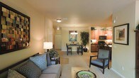 This single-floor residence offers an open-concept layout that seamlessly connects the main living spaces, creating a cohesive and functional environment. The living area is warmly lit and comfortably furnished, featuring plush seating with patterned accent pillows and soft carpeting underfoot, establishing a relaxed and welcoming ambiance. A decorative table lamp adds a warm glow that complements the neutral, earth-toned walls throughout the space. The dining area sits centrally within the open floor plan, highlighted by an elegant chandelier overhead and accompanied by a dark-wood dining table and chairs. A large decorative plant nearby adds a natural, organic touch to the interior. The kitchen is visible from both the living and dining areas, featuring rich warm-toned cabinetry, dark countertops, and black appliances including a range and microwave. Upper cabinets extend to near the ceiling, maximizing storage capacity. The seamless flow between the living room, dining area, and kitchen makes this space well-suited for both everyday living and entertaining. The consistent warm color palette and thoughtful lighting throughout create a unified, comfortable atmosphere across the entire floor plan.