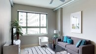 This stylish single-floor urban residence showcases a thoughtfully designed interior with a blend of modern and industrial aesthetic elements. The living area serves as the heart of the space, characterized by large windows that invite an abundance of natural light and frame views of the surrounding city skyline. Exposed ductwork along the ceiling adds an industrial loft-like character, while a ceiling fan contributes both form and function to the room. Neutral gray walls provide a versatile backdrop that complements the contemporary furnishings and vibrant accent colors. A hallway connects the living area to the private quarters of the residence, with clean white trim framing each doorway. The corridor leads to a bedroom featuring the same soft, neutral palette, and a spacious walk-in closet offers generous storage. Track lighting along the hallway ceiling provides practical illumination throughout. Light-toned carpeting in the private areas contrasts pleasantly with the finishes in the main living space, contributing to a warm and cohesive feel. Overall, the space strikes a balance between urban edge and comfortable livability, making it well-suited for those who appreciate contemporary city living.