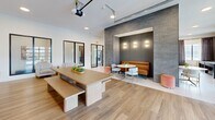 Located at 7298 Kensington Way in McCordsville, Indiana, this thoughtfully designed single-floor community space offers a generous and versatile open-concept layout. The interior showcases a contemporary aesthetic defined by warm wood-look flooring, crisp white walls, and bold architectural accents including a dramatic grey textured brick feature wall that anchors a sleek linear fireplace with a wall-mounted television above.

The living area is bright and welcoming, filled with natural light from large black-framed windows dressed with elegant draping curtains that frame views of the surrounding neighborhood. A comfortable crimson sectional sofa and area rug define a relaxed lounge zone, while a dedicated game area with a full shuffleboard table adds a recreational dimension to the space.

A cozy built-in banquette nook, finished with textured grey tile surround and warm sconce lighting, provides an intimate seating area alongside a communal dining table with bench seating — ideal for gathering and socializing. The space also features access to an adjacent kitchen area with stainless steel appliances and rich cabinetry, accented by a deep green feature wall that adds a pop of color. Recessed lighting and a track-mounted projector system further enhance the space's functional and entertaining appeal.