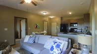 This single-floor space offers a thoughtfully designed open-concept layout that integrates the living, dining, and kitchen areas into one cohesive and functional environment. The living room serves as the heart of the space, featuring a generous L-shaped sofa adorned with decorative throw pillows and a ceiling fan that enhances comfort throughout the year. Warm neutral wall tones create a welcoming ambiance, complemented by recessed lighting and flush-mount ceiling fixtures that ensure the space is well-lit. A vibrant piece of wall art adds a pop of color and personality to the room. The dining area is situated adjacent to the living room, offering a compact yet practical table and chair set ideal for casual meals. The kitchen is open to the main living area via a breakfast bar counter, providing additional seating and a natural gathering point. It is equipped with dark appliances, including a refrigerator, microwave, and stove, alongside warm-toned cabinetry that adds a classic touch. Multiple interior doors provide access to additional rooms, suggesting a well-organized and versatile floor plan. Overall, the space presents a comfortable and practical layout suited for everyday living.