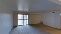 This single-floor residence offers a clean, functional layout designed for comfortable everyday living. The main living area is generously sized, featuring wall-to-wall carpeting that adds warmth underfoot and a soft, neutral tone that complements the light beige walls. A large sliding glass door serves as the focal point of the living space, flooding the room with natural light and offering views of the surrounding greenery outside. Vertical blinds frame the sliding door, providing both privacy and light control as needed. The textured ceiling adds a subtle architectural detail, while a floor-level vent indicates a built-in climate control system for year-round comfort. The open wall space throughout the living area provides flexibility for a variety of furniture arrangements, making it easy to personalize the room. The overall ambiance is bright, airy, and move-in ready, with a straightforward design that prioritizes open space and natural light. The neutral finishes throughout create a versatile backdrop well-suited to a range of interior styles and décor preferences.