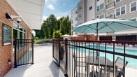 Located at 2940 Falstaff Rd in Raleigh, NC, this contemporary multi-story residential building presents a well-designed exterior and a standout outdoor amenity area that serves as a central gathering space. The building's façade combines neutral-toned siding, brick accents, and large windows across three floors, offering a clean and modern aesthetic that blends cohesively with the surrounding landscape.

The outdoor pool area is a defining feature of this property. A generously sized, crystal-clear swimming pool anchors the courtyard-style space, with easy-entry steps and a wide surrounding deck finished in light-toned concrete. Teal-cushioned lounge chairs and chaise lounges are thoughtfully arranged for relaxation, while multiple patio umbrella stations in both teal and natural tones provide shaded seating with bistro-style tables and chairs, ideal for outdoor dining or socializing.

The pool area is enclosed by sleek black metal fencing, providing both security and an open sightline to the mature trees and greenery that border the property. The building wraps around a portion of the courtyard, creating a semi-enclosed atmosphere that feels private yet connected to the outdoors. Ground-level interior spaces with large glass windows face the pool, enhancing indoor-outdoor connectivity. The overall ambiance is resort-like, combining practical amenities with an inviting, well-maintained outdoor environment.