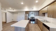 This single-floor residence showcases a contemporary design with a cohesive aesthetic carried throughout its living spaces. Wood-look plank flooring runs continuously across the unit, complementing the warm-toned cabinetry found in both the kitchen and bathroom.

The kitchen serves as a functional centerpiece, featuring an L-shaped layout with medium wood-grain cabinetry extending to the upper walls for generous storage capacity. White quartz-style countertops provide a clean contrast against the cabinetry, while a full suite of stainless steel appliances — including a refrigerator, electric range with overhead exhaust, and dishwasher — ensures modern convenience. A freestanding kitchen island with a matching white countertop and additional storage offers extra workspace and casual dining potential.

The kitchen opens directly into the living area, which benefits from a ceiling fan, a glazed exterior door, and a window fitted with horizontal blinds, allowing natural light to fill the space. A separate bedroom features its own ceiling fan and window with outdoor views.

The bathroom is cleanly finished in white, with a soaking tub and shower combination equipped with grab bars, a toilet, and a wood-grain vanity with a white countertop, all continuing the unit's consistent modern palette.