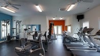 Located at 500 Talon Dr in Jacksonville, NC, this fitness center offers a well-appointed workout space designed to support a comprehensive exercise routine. The single-floor facility is bright and open, illuminated by overhead fluorescent lighting that keeps the space feeling energized and functional throughout the day. A bold orange accent wall serves as a striking focal point at the far end of the room, adding a sense of motivation and energy to the environment. Large windows and glass-paneled doors allow natural light to filter in, contributing to an open and inviting atmosphere. The flooring features durable, dark rubberized material well-suited for high-traffic fitness use. Cardio equipment, including stationary bikes, elliptical machines, and treadmills, is thoughtfully arranged to maximize floor space and ease of movement between stations. A multi-station cable weight machine and a full dumbbell rack provide ample options for strength training. Floor-to-ceiling mirrors along one wall enhance the sense of space and allow users to monitor their form. The layout is clean and organized, making the facility accessible and easy to navigate for users of all fitness levels.