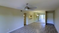 This single-floor space offers an open and airy layout, thoughtfully designed to maximize both functionality and comfort. The living area is anchored by smooth, neutral-toned walls and features stylish hardwood-style flooring that extends throughout, creating a cohesive and welcoming atmosphere. A modern ceiling fan with an integrated light fixture serves as a subtle focal point overhead, providing both style and practicality.

A large mirrored wall runs along one side of the living space, visually expanding the room and reflecting natural light that filters in through vertical blinds covering a sliding glass door, hinting at an outdoor area beyond. This design element adds depth and brightness to the interior.

The space flows naturally into a kitchen area, partially visible through a pass-through opening in the wall. The kitchen features pendant lighting and counter seating, making it well-suited for both casual dining and entertaining. Additional built-in shelving is accessible through a nearby alcove, offering practical storage solutions.

Throughout, the clean lines, neutral color palette, and consistent flooring create a unified and inviting environment. The combination of natural light, reflective surfaces, and an open floor plan gives this space a sense of spaciousness and ease, making it a versatile and functional living environment.
