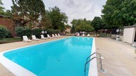 This outdoor pool area offers a generously sized rectangular swimming pool filled with clear, vibrant blue water. The pool is framed by a smooth concrete deck that provides ample space for lounging, lined with a row of white plastic lounge chairs positioned along one side—ideal for relaxing poolside. Stainless steel handrails are installed at the pool's entry point, ensuring safe and convenient access to the water. Lane markers are visible along the pool's surface, suggesting the space accommodates both recreational and lap swimming. The pool is enclosed by a chain-link fence that defines the perimeter and provides a layer of security. Informational and safety signage is posted near the entry gate. The surrounding landscape features a mature canopy of tall trees, including evergreens and deciduous varieties, creating a shaded, park-like atmosphere that enhances the outdoor experience. The overall setting is open and well-maintained, offering a communal recreational space designed for warm-weather use.