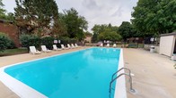 This outdoor pool area offers a generously sized rectangular swimming pool filled with clear, vibrant blue water. The pool is framed by a smooth concrete deck that provides ample space for lounging, lined with a row of white plastic lounge chairs positioned along one side—ideal for relaxing poolside. Stainless steel handrails are installed at the pool's entry point, ensuring safe and convenient access to the water. Lane markers are visible along the pool's surface, suggesting the space accommodates both recreational and lap swimming. The pool is enclosed by a chain-link fence that defines the perimeter and provides a layer of security. Informational and safety signage is posted near the entry gate. The surrounding landscape features a mature canopy of tall trees, including evergreens and deciduous varieties, creating a shaded, park-like atmosphere that enhances the outdoor experience. The overall setting is open and well-maintained, offering a communal recreational space designed for warm-weather use.