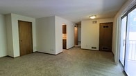 This single-floor residential space offers a straightforward and functional layout designed for comfortable everyday living. The main living area is generously sized, featuring wall-to-wall carpeting in a neutral tone that adds warmth underfoot and contributes to the overall subdued, clean aesthetic. Crisp white walls and flush-mount ceiling lighting keep the space feeling bright and open.

A partial dividing wall separates the living area from the kitchen zone, providing a sense of definition between spaces while maintaining an open feel. The kitchen features dark wood cabinetry that contrasts with the lighter walls, offering ample storage. The cabinetry design is classic and utilitarian, complemented by a light-colored countertop visible in the background.

Multiple doorways lead to additional rooms throughout the unit, suggesting a well-connected floor plan. Dark-stained wooden door frames and baseboards run consistently throughout, providing a cohesive visual detail that ties the interior together. HVAC floor vents are present in the main living area, ensuring climate control throughout the space.

Overall, the space presents a clean, neutral, and move-in-ready environment with a practical design that prioritizes functionality and ease of use across its single level.