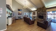 This single-story clubhouse is designed to serve as a welcoming community gathering space, combining comfort, functionality, and architectural character throughout. The great room immediately impresses with soaring vaulted ceilings supported by exposed white-painted beams, creating an airy and open atmosphere. Warm hardwood flooring runs throughout, complementing the space's inviting aesthetic.

A floor-to-ceiling stone fireplace serves as a dramatic focal point in the main lounge area, where plush leather sofas and a glass-top coffee table are arranged for relaxed socializing. An elegant chandelier anchors the space, while a wall-mounted television adds a modern amenity. French doors and large sliding glass windows allow abundant natural light to pour in and provide views of the surrounding outdoor greenery.

The billiards area features a full-size pool table beneath pendant lighting, offering an entertaining option alongside a dedicated dining section with wooden tables and chairs. A kitchenette with light-toned cabinetry, a breakfast bar with seating, and stainless steel appliances supports catering and event needs.

A dining room with floor-to-ceiling windows and sliding glass door access leads to an outdoor deck, where patio seating beneath umbrellas overlooks a landscaped exterior. Throughout the clubhouse, the open layout and abundance of natural light create a bright, social, and versatile environment suited for a wide range of community activities.