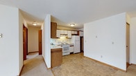 This single-floor residence offers a practical and well-organized layout designed for comfortable everyday living. The kitchen is a standout feature, showcasing a combination of white upper cabinets and warm wood-toned lower cabinetry that provides ample storage space. White appliances, including a refrigerator, electric range, and range hood, are neatly integrated into the kitchen's layout, which also includes a double sink and open shelving for added convenience. The kitchen transitions seamlessly into the adjacent living area, creating an open-concept feel that enhances the sense of space. The flooring shifts from resilient tile in the kitchen to soft carpet in the living areas, offering a comfortable underfoot experience throughout. Neutral wall tones and white ceilings keep the interior feeling bright and clean, while flush-mount ceiling light fixtures provide consistent illumination across the space. A hallway visible from the main area leads to additional rooms, suggesting a thoughtful and connected floor plan. The overall ambiance is clean, functional, and move-in ready, making it well-suited for those seeking a straightforward and dependable living space.