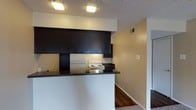 This single-floor residence offers a practical and functional layout designed for comfortable everyday living. The kitchen serves as a central feature of the space, showcasing dark espresso-toned upper and lower cabinetry that creates a bold contrast against the lighter walls. A peninsula-style counter with a dark granite-look surface extends into the living area, providing additional workspace and a casual dining or seating option. The kitchen is equipped with a white electric range and a refrigerator, offering everything needed for home cooking. Recessed flush-mount ceiling lights illuminate the kitchen area, highlighting the textured ceiling overhead. A HVAC vent is visible along the upper wall, indicating a central air and heating system throughout the unit. The flooring transitions from warm hardwood-style planks in the kitchen and hallway areas to carpeting in the adjacent living spaces, adding texture and comfort underfoot. Neutral, warm-toned walls throughout contribute to an inviting ambiance. The open sightlines between the kitchen and adjoining hallway create a sense of connectivity across the floor plan, making the space feel open and easy to navigate.