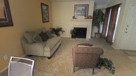 This single-floor residence offers a comfortable and inviting atmosphere anchored by a well-appointed living room. The space centers around a classic tile-surround fireplace, complete with a wood mantel that serves as a natural focal point — adorned with framed artwork and decorative accents that add a personal, homey touch. Warm, neutral-toned walls complement the plush wall-to-wall carpeting, creating a soft and cohesive aesthetic throughout. Seating is generously arranged, featuring a cushioned sofa with decorative pillows alongside a patterned accent chair, offering ample space for relaxation and conversation. Framed landscape artwork punctuates the walls, contributing to the room's warm character. Lush indoor plants are thoughtfully placed throughout the living area, bringing natural life and vibrancy to the interior. Drapery along the windows hints at natural light potential, while the overall layout is open and functional. The single-floor design ensures easy accessibility and a seamless flow throughout the home. The overall ambiance is warm, grounded, and lived-in — ideal for everyday comfort and casual entertaining.