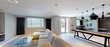 This single-floor amenity space is thoughtfully designed with a contemporary aesthetic, combining multiple functional zones within an open, cohesive layout finished in neutral tones, large-format tile flooring, and recessed lighting throughout.

The lounge and media area anchors one end of the space, featuring a generously sized sectional sofa, an accent armchair, and a glass coffee table arranged on a textured area rug. A large wall-mounted flat-screen television is set against a dark wood-paneled feature wall, complemented by framed abstract artwork, creating a polished, relaxed atmosphere.

The open central area transitions seamlessly into a game and dining zone, where a long communal game table sits beneath a cluster of striking black pendant lights. Adjacent seating with high-top stools lines the perimeter near large windows, allowing natural light to fill the space and providing outdoor views.

The kitchen area is equipped with a central island featuring bar-height seating for multiple guests, warm wood cabinetry, a mosaic tile backsplash, built-in televisions, and ample counter space — well-suited for catering or social gatherings.

The dining nook offers several café-style tables and chairs beside floor-to-ceiling windows, brightened by natural light and accented with colorful artwork, completing this versatile and well-appointed communal space.