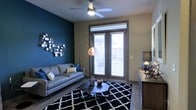 This single-floor space makes a striking first impression with its bold design choices and contemporary aesthetic. The living area serves as the centerpiece, anchored by a deep teal accent wall that adds dramatic contrast against the neutral surrounding walls. A decorative metallic wall art piece enhances the visual interest of this feature wall, lending the space an artistic, curated feel.

The room is furnished with a sleek gray sofa paired with coordinating throw pillows, positioned over a bold black-and-white geometric area rug that grounds the seating arrangement. A low-profile, multi-piece coffee table sits at the center, offering both style and function. A sculptural arc floor lamp with a copper-toned shade provides warm, ambient lighting that complements the ceiling fan and light fixture overhead.

Natural light flows into the space through a set of glass French doors fitted with horizontal blinds, offering both privacy and access to an outdoor area beyond. The hardwood-style flooring runs throughout, adding warmth and continuity to the space. A media console along the opposite wall provides additional storage and display surface, completing the room's modern, well-appointed layout. Overall, the space exudes a contemporary urban character with thoughtful design details throughout.