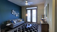 This single-floor space makes a striking first impression with its bold design choices and contemporary aesthetic. The living area serves as the centerpiece, anchored by a deep teal accent wall that adds dramatic contrast against the neutral surrounding walls. A decorative metallic wall art piece enhances the visual interest of this feature wall, lending the space an artistic, curated feel.

The room is furnished with a sleek gray sofa paired with coordinating throw pillows, positioned over a bold black-and-white geometric area rug that grounds the seating arrangement. A low-profile, multi-piece coffee table sits at the center, offering both style and function. A sculptural arc floor lamp with a copper-toned shade provides warm, ambient lighting that complements the ceiling fan and light fixture overhead.

Natural light flows into the space through a set of glass French doors fitted with horizontal blinds, offering both privacy and access to an outdoor area beyond. The hardwood-style flooring runs throughout, adding warmth and continuity to the space. A media console along the opposite wall provides additional storage and display surface, completing the room's modern, well-appointed layout. Overall, the space exudes a contemporary urban character with thoughtful design details throughout.