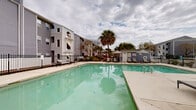 This contemporary residential complex offers a blend of comfort and leisure. The centerpiece is a spacious, resort-style swimming pool with a generous deck area, perfect for sunbathing and relaxation. The pool is surrounded by comfortable lounge chairs and umbrellas, providing ample seating options for residents.

The architecture is characterized by multi-story buildings with a mix of gray and white exterior finishes, creating a clean and modern aesthetic. Large windows and balconies offer residents outdoor spaces and natural light. Distinctive arched windows on the upper floors add architectural interest.

Landscaping plays a significant role in the complex's ambiance, with mature palm trees dotting the grounds, lending a tropical feel to the environment. Well-maintained shrubbery and green spaces further enhance the property's appeal.

For outdoor entertaining, the complex features communal seating areas with modern patio furniture, ideal for socializing or enjoying a meal al fresco. A perimeter fence ensures privacy and security for residents.

The property appears to offer additional amenities, possibly including a clubhouse or communal building, as seen in the images. Overall, this apartment complex presents an attractive living space with a focus on outdoor enjoyment and community living.