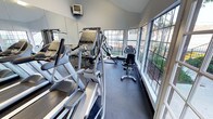 This fitness center is thoughtfully designed to support a well-rounded workout routine within a bright, open environment. The space benefits from vaulted ceilings that lend an airy, expansive feel, while large multi-pane windows line one wall, flooding the interior with natural light and offering pleasant views of surrounding greenery.

The cardio section is prominently featured and includes multiple treadmills, elliptical trainers, a stair climber, and a stationary recumbent bike, providing ample options for aerobic exercise. A wall-mounted television is positioned for convenient viewing during cardio sessions. Strength training is also accommodated, with a cable machine and additional resistance equipment visible along one side of the room.

Full-length mirrors span a significant portion of the wall space, enhancing the sense of depth while allowing users to monitor their form during workouts. The flooring is a durable, low-pile carpet suited for fitness use, and recessed and overhead lighting supplements the natural light throughout the day. The overall ambiance is clean, functional, and well-maintained, making it a practical and inviting space for daily exercise.