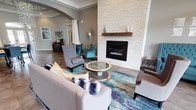 This well-appointed single-floor community space combines sophisticated design with functional layout across several distinct areas. The main lounge is a standout, anchored by a floor-to-ceiling stacked white stone fireplace with a rich wood mantel, creating a warm and inviting focal point. Comfortable seating arrangements — including tufted wingback chairs in deep teal and neutral upholstered sofas — are accented by a vibrant area rug in blues and golds, lending the space a contemporary, curated feel. Tall ceilings with crown molding and recessed lighting elevate the room's sense of openness, while large French doors with transom windows allow natural light to pour in. A separate seating nook with teal accent chairs and a sunburst wall mirror adds an intimate gathering area. An adjacent dining space features a round high-top table beneath a striking orb chandelier set within a coffered ceiling detail. A service window connects to a back office area, indicating a professional leasing or administrative function. The kitchen is equipped with white shaker cabinetry, black granite countertops, and a full suite of black appliances, offering a practical and polished culinary space. White architectural columns and arched passageways define zones throughout, contributing to the space's refined, cohesive aesthetic.