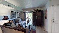 This single-floor residence centers around a thoughtfully designed living room that balances comfort and style. The space is anchored by a plush gray sofa adorned with patterned throw pillows, complemented by additional seating that creates an inviting conversational arrangement. A round glass-topped coffee table serves as a functional centerpiece, topped with a decorative floral arrangement that adds a natural touch.

Natural light flows generously into the room through sliding glass doors that open to a private balcony, offering views of lush greenery beyond. Sheer curtains frame the doors, softening the light while maintaining an airy feel. The neutral-toned walls provide a versatile backdrop, accented by a pair of framed botanical artwork pieces that add visual interest.

A large, dark wood armoire serves as a striking focal point on one wall, providing substantial storage while contributing warmth and contrast to the lighter tones of the room. Decorative items displayed atop it add a personalized touch. The flooring transitions between a soft area rug anchoring the seating area and light hardwood-style flooring extending toward the perimeter, enhancing the overall sense of warmth and cohesion throughout the space.