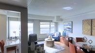 This single-floor space is centered around a thoughtfully designed living area that balances comfort and contemporary style. Large windows span the exterior wall, flooding the room with abundant natural light and offering expansive views of the surrounding landscape and skyline, making the space feel open and airy. The textured ceiling adds subtle character overhead, while warm hardwood flooring grounds the room with a sense of warmth and continuity.

The living area is furnished with a coordinated arrangement that includes a plush grey sofa, a charming blue accent armchair with decorative pillows, and a distinctive round mosaic coffee table atop a patterned area rug. A stylish table lamp provides additional ambient lighting, complementing the natural light from the windows. A wooden console with ornate detailing serves as both a functional and decorative piece, topped with sculptural blue vases that echo the cool tones found throughout the room. A bold abstract artwork anchors one wall, adding a vibrant focal point. The overall ambiance is bright, curated, and inviting, with a cohesive color palette of blues, greys, and warm wood tones.