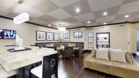 Located in Dallas, this well-appointed single-floor lounge space combines sophistication with comfort through thoughtful design and premium finishes. The room is anchored by a striking checkered ceiling with recessed lighting and a central pendant light fixture, adding visual depth and a refined ambiance throughout.

Rich, dark hardwood flooring runs the length of the space, providing a warm contrast to the cream-toned walls adorned with a curated selection of framed artwork. The layout is divided into distinct seating zones, offering flexibility for both social gatherings and relaxed individual use. A banquette-style seating arrangement with upholstered booth benches and wooden tables creates a cozy dining or meeting area, while a plush, gold-toned sofa with chevron accent pillows provides a more casual lounge setting.

A wall-mounted flat-screen television serves as a focal point in the seating area, enhancing the space's functionality as an entertainment hub. The kitchen bar area features a stunning marble-topped counter with dark cabinetry and sleek bar seating, ideal for casual dining or socializing. The overall design palette—neutral tones, dark wood accents, and metallic details—creates an inviting and polished atmosphere well-suited for community gatherings or leisure.