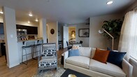 This single-floor residence offers a thoughtfully designed open-concept layout that seamlessly connects the living, dining, and kitchen areas into one cohesive and functional space. The living area serves as the heart of the unit, furnished with a comfortable white sofa accented by colorful throw pillows and complementary accent chairs, all anchored by a patterned area rug atop warm wood-look flooring that extends throughout the space.

The kitchen is positioned to the left of the main living area and features dark cabinetry, light stone countertops, and a raised breakfast bar with industrial-style metal bar stools — ideal for casual dining or entertaining. A compact dining area with a table and chairs sits just beyond the living space, offering a dedicated zone for meals.

A doorway visible from the main living area provides a glimpse into the bedroom, which is warmly lit and accented with decorative touches. The overall color palette is neutral and modern, with warm beige and gray tones on the walls complemented by pops of rust, blue, and earthy accents in the furnishings. Recessed lighting and structured ceiling details add to the clean, contemporary ambiance of the space.