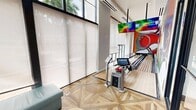 This striking single-floor entertainment space is designed as a high-end private bowling alley, offering a seamless blend of sport and sophisticated interior design. The centerpiece is a full-length bowling lane finished in classic wood, flanked by glass guardrails and anchored at the far end by a bold, colorful mural depicting oversized bowling balls and abstract graphic forms in vivid reds, greens, and blues. Large-format display screens are mounted overhead above the lane, providing a dynamic visual element for scorekeeping and entertainment. The lounge area adjacent to the lane features teal sectional seating accented with orange and patterned throw pillows, arranged around a gold accent table — offering a comfortable viewing and gathering space. The flooring throughout the lounge area is a striking geometric parquet wood pattern that adds warmth and visual texture. Floor-to-ceiling windows dressed with sheer roller shades line one wall, flooding the space with abundant natural light and offering views of lush tropical palm trees outside. Black-framed glass partitions and walls connect the bowling area to adjacent spaces, maintaining an open, airy feel while defining the room. The overall ambiance is energetic yet refined, making this an exceptional private entertainment venue.