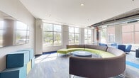 This generously sized single-floor amenity space offers a dynamic and well-appointed environment designed for community engagement and recreation. At the entrance, a modern reception desk anchored by bold "sb" branding sets a welcoming tone, with elevator access and mailboxes conveniently nearby.

The open lounge area is a standout feature, centered around a distinctive circular sectional sofa in gray and lime green with a round coffee table at its core. Large windows flood the space with abundant natural light and provide views of the surrounding outdoor greenery. Additional seating clusters with blue and charcoal armchairs create versatile social zones throughout, accented by colorful artwork and vibrant patterned carpeting in blue, gray, and yellow tones.

The game room is comprehensively outfitted with a foosball table, full-size billiards table, shuffleboard table, and ping-pong table, all beneath stylish pendant lighting. Wall-mounted flat-screen TVs and high-top bistro tables add to the entertainment-focused atmosphere.

Throughout the space, exposed ductwork and concrete structural elements lend an industrial-modern character, complemented by a bright, contemporary color palette. The overall design balances energy and comfort, making it an ideal communal hub for socializing and recreation.