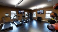 This fitness center is designed to provide a comprehensive workout environment within a compact, single-floor layout. The room features dark hardwood flooring that lends a clean, polished aesthetic throughout the space. Warm earth-toned walls create an energizing atmosphere, complemented by bright overhead fluorescent lighting that ensures excellent visibility across the entire floor.

The cardio section is equipped with a treadmill and an elliptical trainer, positioned near the windows to allow natural light and outdoor views during workouts. A multi-functional cable resistance machine occupies the center of the space, offering a wide variety of strength training exercises. A dedicated free weight area includes a tiered dumbbell rack stocked with multiple weight increments, supporting both beginner and advanced training routines.

Multiple large flat-screen TVs are mounted on the walls, providing entertainment options for users during their sessions. Several windows are evenly distributed along the walls, inviting natural light and giving the room an open, airy feel. The layout is thoughtfully organized to maximize usable floor space, leaving ample room for stretching or floor exercises. Overall, this fitness center offers a well-rounded, functional, and visually appealing environment suited to a variety of fitness needs.