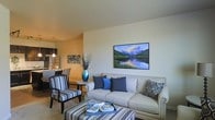 This single-floor space offers a thoughtfully designed open-concept layout that blends comfort and style. The living area serves as the heart of the home, centered around a cream-toned sofa accented with navy blue throw pillows and complemented by a coordinating striped armchair and ottoman. A large landscape artwork depicting a serene mountain lake adds a striking focal point to the neutral wall. Natural warm lighting enhances the inviting ambiance throughout. The living space flows effortlessly into a dining area, where a small round side table and chairs create an intimate setting. Just beyond, the kitchen is visible with dark cabinetry, stainless steel appliances — including a refrigerator — and pendant lighting that adds a modern touch overhead. The flooring transitions smoothly between the spaces, reinforcing the cohesive, open feel of the layout. Decorative elements such as a blue vase with tall branches and layered textiles add personality and warmth. The overall design palette of navy, cream, and warm neutrals creates a calm, polished atmosphere that is both functional and aesthetically refined.