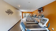 Located at 300 20th Ave W in Milan, Illinois, this single-room fitness center is a well-equipped and efficiently designed exercise facility. The space features bold orange accent walls that energize the environment, complemented by neutral beige walls adorned with fitness-themed motivational word art. Recessed lighting and large windows along multiple walls flood the interior with natural light, while a set of French doors provides convenient access.

The cardio section is well-stocked with multiple treadmills, an elliptical trainer, and a recumbent bike, all arranged in a row for easy use. A wall-mounted flat-screen television is positioned above the cardio equipment, offering entertainment during workouts. A full-length mirror spans one wall, visually expanding the space and aiding users with form and technique.

The strength training area includes a multi-tiered dumbbell rack with a variety of weights and an adjustable workout bench, providing options for a range of resistance exercises. The flooring throughout is a durable, speckled rubber-style surface suited for high-traffic fitness use. The space is completed with a fire extinguisher and safety signage, underscoring a commitment to user safety. Overall, this fitness center delivers a functional and motivating workout environment in a compact, thoughtfully arranged layout.