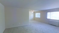 This single-floor space presents a clean, understated aesthetic defined by its open layout and soft, neutral color palette. The main living area is finished with wall-to-wall light beige carpeting that lends a sense of warmth and comfort underfoot. Smooth white ceilings and cream-toned walls create a bright, airy atmosphere throughout the space.

A ceiling fan is centrally positioned in the main area, providing both practical air circulation and a subtle design element. A window along one wall allows natural light to filter into the room, further enhancing the open and welcoming feel of the interior. The clean lines of the walls and the absence of visual clutter give the space a versatile, move-in-ready character that can easily accommodate a variety of furnishing styles.

The layout makes efficient use of the available area, with a defined alcove or transitional zone visible from the main room, adding a degree of spatial variety to the otherwise open floor plan. White baseboards provide a crisp contrast to the carpeted floors, completing the tidy, well-maintained look of the interior. Overall, this single-level space offers a calm and functional environment well-suited for comfortable everyday living.