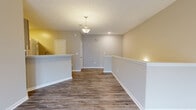 Located at 901 Carlton Park Dr in Flowood, Mississippi, this two-story, one-bedroom, one-bathroom unit offers a well-organized layout spread across two levels. The ground floor houses an attached single-car garage with a concrete floor and automatic opener, providing secure, convenient parking and storage. Carpeted stairs with a white handrail lead up to the main living level, which contains the majority of the living space. The open-concept kitchen and living area forms the heart of the home, featuring rich wood-look vinyl plank flooring, neutral warm-toned walls, and white wainscoting details. The kitchen is well-appointed with gray shaker-style cabinetry, a subway tile backsplash, light quartz countertops, and a full suite of stainless steel appliances including a gas range, built-in microwave, dishwasher, and a French door refrigerator. A pendant light fixture illuminates the kitchen bar area, adding a polished touch. The spacious bedroom offers plush carpeting, natural light from a window with blinds, and an arched entryway detail. The full bathroom includes a tub/shower combo with white tile surround, a quartz-topped vanity with gray cabinetry, and wood-look flooring, rounding out a clean and functional space.